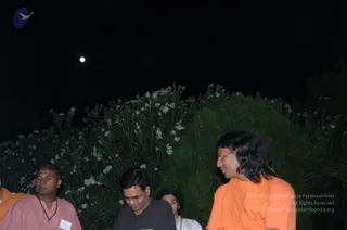 Night photograph outdoors beside a dense hedge of white-flowered oleander under a dark sky with a moon. On the right, a man with long dark hair wears an orange saffron robe with a wrap over the shoulder, appearing to be a person candidate by appearance cues of long swept hair and saffron clothing. Beside him, a bespectacled man in a dark t-shirt looks downward. At left, a man in a pink kurta wears a rudraksha-style mala and a white name badge. Behind them, a person in white with a red mala is partly visible.