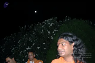 A night photograph outdoors beneath a dark sky with a bright moon at upper centre. Dense shrubs with small white flowers and green foliage fill the background. In the foreground at right, a man with long, wavy black hair wears an orange garment and has a red bindi-style mark on his forehead; the figure is partly blurred. Behind him, a man with glasses wears a white shirt and an orange shawl, and at far left a partly visible man in dark clothing stands near the bottom edge. a person cues include long swept-back hair, orange clothing and a forehead mark, but identity is uncertain from appearance.