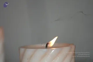 Close-up photograph of a single lit white pillar candle with a twisted, ribbed surface in pale cream and peach tones. A small flame burns from a dark wick, with thin wisps of smoke curling upward against a plain grey wall. The background is blurred, with another candle partly visible at the left edge. No people are visible.