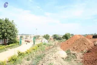 A dusty construction site on a rural road with a pale sandy track running past two green gates on the left. A single worker in light clothing walks on the track near a red truck parked beyond the gates. On the right, large mounds of reddish-brown earth and grey soil pile up beside a ditch with shrubs and a large grey boulder. Tall lamp posts, scattered trees and low buildings line the open, hazy landscape under a pale blue sky with wispy clouds.