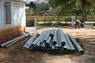 A construction site photograph showing a large stack of grey PVC pipes of varying diameters lying on bare red-brown earth beside a white-washed concrete building with a small window. A palm tree and a large boulder sit behind the pipes. In the background, two workers in white and light clothing, one in a white shirt and one in orange-brown trousers, are working near a low wall, while a third person in a teal top crouches on the right. Loose stones and a wooden rail are scattered across the ground.