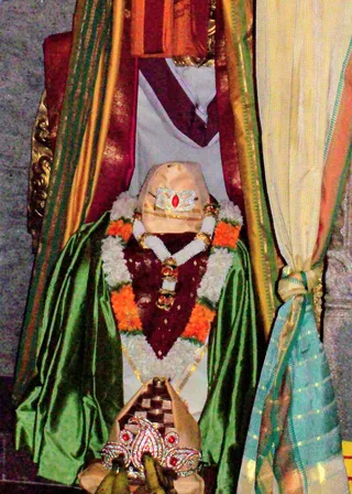 A photograph of a decorated deity murti seated within a draped alcove. The figure wears a green silk garment over a maroon velvet vest, with garlands of white flowers and orange marigolds around the neck and shoulders. A silver crown with a red central jewel sits atop the head, and a shaped stone or metal pedestal in front bears a jewelled ornament. Red and orange drapes frame the niche, with a cream and teal cloth tied at the right.
