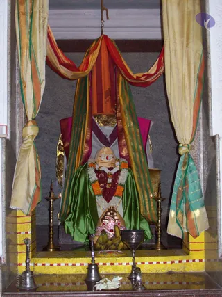 A photograph of a shrine niche with draped fabric in orange, red, green and cream with gold borders framing a seated figure. The figure is a cloth-draped form with a gold and jewelled headpiece, wearing a garland of white jasmine and orange marigold and a red-and-white beaded necklace, with a green satin cloth draped over the lower body. Before it stands a silver-toned bell and a brass vessel with a white flower offering, brass oil lamps flank the seat, and bananas and a jewelled tray sit on the yellow-painted platform. No physical people are visible; the seated figure is a statue or murti, not a visible person.
