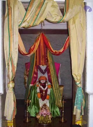 A decorated shrine niche framed by gold-and-cream silk drapes with green and orange borders. Beneath a hanging canopy of red, orange and green cloth sits a seated figure, presented as a deity murti or statue rather than a living person. It wears a green satin cloth, a white and orange flower garland, a red-and-gold ornamental necklace, and an orange cap with a white forehead mark. Brass oil lamps flank the shrine, and in front stand bananas and a jeweled crown-like ornament on a tray. No people are visible.