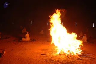 Night photograph of a large open sacred fire burning on bare reddish ground, with tall orange flames rising from a pile of logs at the right of the frame. In the dim background several people in white clothing sit on mats around the fire area, one seated figure in white with a white head wrap facing away from the camera near the left. Faint blue-white lights stand in the background. The fire appears to be part of a fire ritual, consistent with a homa or agni process. No identifiable a person depiction is visible.