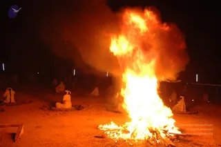 Night photograph of a large open fire blazing on bare reddish ground, with thick smoke rising and drifting left. Several people in white clothes sit cross-legged on mats and cushions around the fire at a distance, some with white head wraps. Small lights stand in the background. The sacred fire ritual setting suggests a homa or agni process.