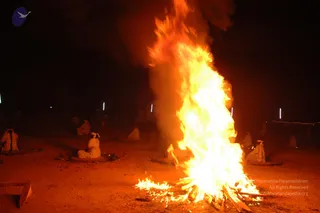 Night photograph of a large sacred fire blazing tall and bright on a dusty open ground. Several people in white clothes sit cross-legged on mats around the fire at a distance, some with heads bowed. Dark smoke rises above the flames. Small lights mark the edges of the ground in the dark background. The scene suggests a fire ritual in progress.