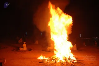 A night photograph of a large, tall bonfire blazing on bare reddish ground, with thick smoke rising behind it. Several people in white clothes sit cross-legged around the fire at a distance, some with white head cloths, while many more figures sit in the dim background. Smaller flames burn at the base of the main fire among logs. The scene suggests a fire ritual, though the precise rite is not clear from the image.