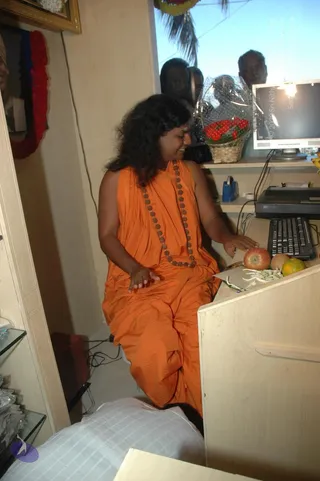 A photograph of a shop interior with a seated figure in an orange-saffron sleeveless robe draped over the body and a rudraksha mala of brown beads around the neck. The figure has long curly dark hair and a vertical mark on the forehead, and sits at a light wooden counter touching a red apple and a yellow fruit, with a computer monitor and keyboard nearby. Behind the counter a framed portrait hangs on the wall with a red and blue garland, and a wrapped basket of red flowers sits on a shelf. Two or three people stand outside the window in the background. Glass display shelves hold packaged goods at left.