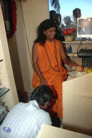 A photograph inside a small shop or office. A long-haired man in a sleeveless orange wrap and a beaded rudraksha mala sits at a wooden counter with a computer monitor and keyboard, smiling, with a red apple and fruit on the counter. A man in a white checked shirt bows low with his head near the seated man's knee. Red flower garlands hang on the left wall and a flower basket sits on a shelf behind the counter. Partially visible people stand in the reflection in the window behind. Swamiji (probable): the seated man has long swept-back hair, an orange wrap and a rudraksha mala, cues consistent with the page's reference to Swamiji.