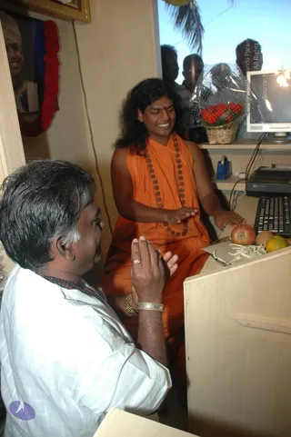 Photograph of a small office interior. A long-haired man in a saffron-orange sleeveless robe with a rudraksha mala and a red tilak sits at a wooden counter, palms hovering over it with a gentle gesture. Opposite him a man in a white shirt with a rudraksha mala and silver bracelets sits with palms pressed together in greeting. On the counter sit a computer keyboard, an apple, an orange, a red-and-white fruit and a basket of red flowers wrapped in cellophane. Behind them a framed portrait with a red garland hangs on the wall, and three onlookers stand near a window. Probable Swamiji (long swept-back hair, saffron robe, rudraksha mala) is seated in the saffron robe at the counter.