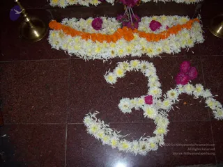 Floor decoration on dark red-brown granite tiles. An Om symbol is formed from white and yellow chrysanthemum-type flowers with scattered pink petals and a few pink blossoms. Above it, a curved band of white flowers edged with orange marigold garlands runs across the top of the frame. Pink flowers rest on the band near a cluster of stems. Brass lamp stands with bell-shaped bases sit at the upper left and right corners. No people are visible.