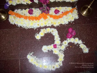 A photograph of floral decoration on a red-and-maroon granite floor. A large Om symbol is formed from white and yellow chrysanthemum petals and blossoms. Above it, a curved band of orange marigold garlands is laid over white flowers, with pink flowers and loose petals scattered across the floor. Brass lamp stands with bowls sit at the top edges of the frame. No people are visible.