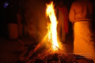 A night photograph of a tall open fire burning on a sandy ground, with dry straw and sticks heaped at its base and flames rising high. Behind the fire, several people in white and cream dhotis and shawls stand in the dim background. On the left, figures in pale garments stand near the dark edge of the frame. A man in white clothing with a red shawl draped over his shoulders stands to the right of the flames, and a blurred figure in a white dhoti occupies the right edge. The scene suggests a homa or yagna fire ritual at night.