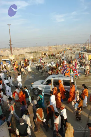 A dusty road at the Allahabad Kumbh Mela crowded with pilgrims, with rows of white tents and makeshift stalls lining the route. At the centre, a silver SUV moves slowly through the crowd, and several figures in saffron-orange robes walk beside it, one holding a multicoloured umbrella with red, green and blue panels. Men in white dhotis and women in saris walk among the throng, and a man with an orange turban and shawl stands near the vehicle. Tall utility poles and power lines stretch across a pale blue sky, with a faint purple logo in the upper left corner.