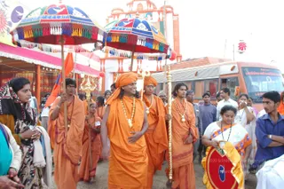 A procession on a dirt road at a Kumbh Mela site. Behind him, another saffron-robed man in a turban and gold necklaces walks, and two young men in orange robes carry tall gold-tipped staffs topped with trident-like finials. Two decorated umbrellas with multicolored tassels are carried overhead. On the left, a woman in a dark shawl watches, and on the right a woman in a white sari with an orange-and-gold border stands among onlookers in casual clothes. Behind them stand a large orange bus and a pink-and-white temple-style structure with a red banner.