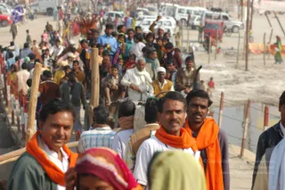 A crowded outdoor procession on a dusty riverside path at the Allahabad Kumbh Mela. Several men in the foreground wear orange saffron shawls over white and green shirts; one has a red tilak on his forehead. A woman in a pink and red headscarf appears at the bottom edge. Behind them a large crowd moves along a bamboo-fenced walkway, including an elderly man in white clothing and a man in a brown vest. Parked white vans and vehicles line the dirt area beside the water, with orange cloth banners on the right.