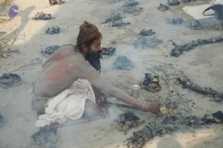 A photograph of a bare-chested sadhu sitting cross-legged on a rug in the open ground, reaching with a long iron rod toward a small brass cup and a metal vessel among scattered charcoal and ash-coloured embers. Smoke rises from the coals, and the man has matted hair tied in a topknot and a long beard, with a white cloth wrapped around his waist. Round soot-blackened fire patches and a folded white mat lie on the sandy ground.