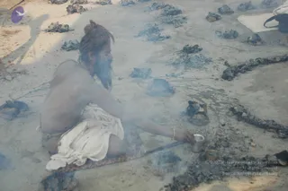 A photograph of a bearded sadhu with long matted hair tied in a topknot, seated bare-chested on a mat in smoky ground dotted with blackened ash mounds and charred logs. He wears a white dhoti and holds a small metal cup while reaching toward the ash, with a long iron rod lying beside him. Smoke drifts across the scene. No ornate a person cues such as saffron robes, crown, or rudraksha are visible, so no candidate is identified.