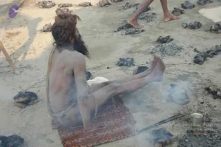 A photograph of a naked ascetic sadhu seated on a patterned mat on bare ground at the Kumbh Mela, viewed from behind. His hair is piled in a topknot and he has a long beard; his body is smeared with grey ash and he wears a thin rope around the waist and shoulders. Scattered dark coal and ash pieces, smoke rising from the ground, a metal cup and a long iron tong lie nearby. Another person's bare feet are visible at the top edge of the frame.