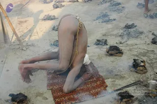 A photograph of a naked ascetic performing a deep forward-bending yoga posture on a woven orange-and-brown mat on bare ground at the Kumbh Mela. His forehead rests on the mat, his buttocks raised, and a thin yellow cord with knots is wrapped around his waist. Scattered blackened cow-dung cakes and smouldering ash lie on the ground around him, with a small metal cup and a pair of sticks at the lower right. Bamboo poles and a cloth tent edge appear at the left. No a person candidate is visible.