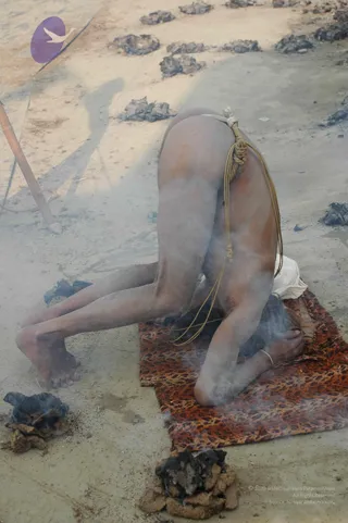 A photograph of a naked ascetic performing a deep forward-bending yoga posture, head bowed to the ground on a brown-and-orange patterned mat on sandy earth. The figure's body is ash-smeared and sun-darkened, with a thin knotted rope or cord wrapped around the waist and a white cloth near the hips. Smoldering cow-dung cakes lie on the ground around the mat, with smoke drifting across the frame.
