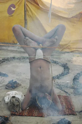 A photograph of an ash-smeared sadhu performing a headstand-style yoga posture on a patterned brown and orange rug at a Kumbh Mela camp. His legs are crossed and raised above him, with his feet interlocked, and he is tied at the waist with a rope that is strung between two lines. Smoke rises from small fires and a ring of burnt ash on the ground around him. Behind him stands a yellow tent with a red emblem, and nearby are a green canvas bag, a folded white cloth, a steel cup and tongs. No a person candidate is visible.
