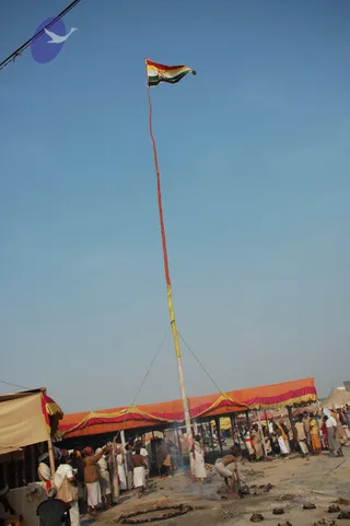 A tall pole with a tricolour-style flag in red, yellow, white and green flies against a clear blue sky at an outdoor encampment. Several men in white dhotis and cloth wraps are raising the pole with ropes, and others stand along an orange and yellow canopied shelter with pink trim. A man bent over near a small fire works on the ground in the foreground, and sandals and scattered bricks lie on the dusty earth. Many people stand in the background among tents.