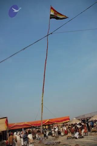 A tall wooden pole wrapped in red and yellow cloth rises from the sandy ground of a Kumbh Mela camp, topped by a triangular flag in red, yellow, white and green stripes with a small figure printed at its centre. Thin guy-lines run from the pole to the ground, and a taut wire crosses the frame above it. Beneath, a crowd of men in white dhotis, saffron and red cloths and bare torsos gathers around the base, several pulling on ropes and lifting or steadying the pole. Behind them an orange and yellow canopy stretches along the camp, with white tents beyond and a clear blue sky overhead. Smoke rises near the base of the pole.