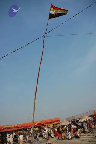 A tall wrapped pole rises diagonally from the lower centre, topped with a triangular flag striped red, yellow, white and green, bearing a small red and gold figure in the centre. Guy wires run to the pole. Beneath it, a crowd of men in white dhotis and orange and red cloth stands on sandy ground, several gripping ropes and lines to raise the pole. Orange and red striped tent awnings line the left side, and white canopy tents stand on the right under a clear blue sky.