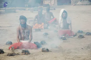 Four sadhus sit cross-legged on sandy ground amid rising smoke from a scattered ring of charred logs, a ritual fire setting. The foremost man, bare-chested with a long beard and white beaded necklace, wears a red bandhani-patterned cloth and sits with hands resting on his lap. Behind him, two men wear orange and red cloths, and a man on the right has a white shawl draped over his head and red lungi, with tilak marks on his forehead. Steel tumblers and small bowls sit near them, with cloth tents and a wooden pole in the background. The scene reads as a fire-side sadhu gathering at the Kumbh Mela.