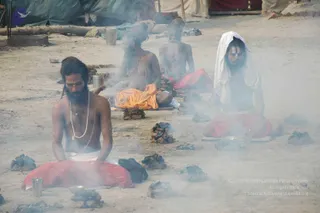 Four sadhus sit on the dusty ground in smoke, surrounded by scattered dung cakes and small metal cups. The man in the foreground wears a red and white dhoti, a white sacred thread and a long pearl mala, with white markings on his bare chest. Beside him, a bearded sadhu in orange cloth sits cross-legged, and another bearded man in red cloth sits behind. At right, a sadhu draped in a white cloth over his head wears a tilak on his forehead and a red dhoti. Tents and a tarpaulin stand behind them, with a tin pot and a log nearby. The scene suggests a sacred fire ritual at the Kumbh Mela.
