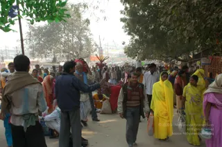 A busy outdoor path at a mela, lined with trees and makeshift stalls on the right. Crowds of pilgrims walk and mingle under hazy winter light. A woman in a bright yellow sari with a shawl over her head walks toward the camera holding a plastic bag, with another woman in yellow beside her. A man in a dark jacket and striped scarf walks in the centre, and an older man in a white dhoti and blue shawl carries a bundle on his head. Women in red, pink and orange saris pass by, and bamboo stalls and flags stand in the background.