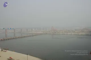 A hazy wide view over a broad, grey river at the Kumbh Mela site on a misty day. A long pontoon bridge of floats with a railing stretches across the water toward the far bank. Along the right, tent camps and a tall red-and-white tower are visible through the fog. A few people sit on the sandy bank at lower left, and a small boat lies near the shore. Banners hang from the steel truss bridge in the background.
