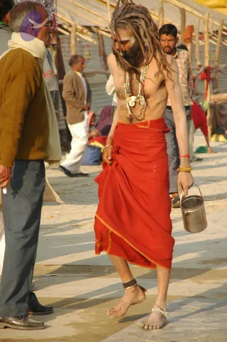 A barefoot sadhu with long matted dreadlocks walks across a sandy riverside path at a crowded Kumbh Mela site. He is bare-chested, wearing a red-orange dhoti with gold-edged border, a multi-strand beaded necklace with shell and metal pendants, and ankle bangles. He carries a stainless steel pot in his right hand. Around him, men in sweaters, jackets and checked shirts stand near temporary bamboo-and-tarpaulin stalls, and a man in a white scarf watches from the left.