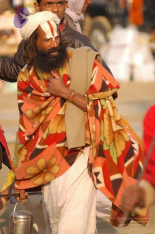A bearded man in a white turban with a white and red vertical tilak mark on his forehead stands outdoors on a sunlit street. He wears a large orange, red and brown floral-patterned blanket wrapped over white dhoti-style clothing, with a beige shawl draped over one shoulder. He holds a metal pot with a handle in one hand and a long rod in the other. Behind him, another man in a dark jacket and a blurred crowd stand near stalls and vehicles.