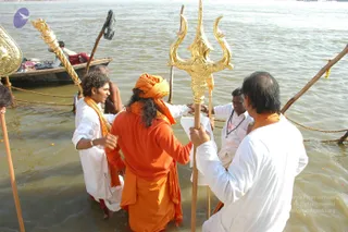 A photograph of people standing waist-deep in a murky river, with a wooden boat and rope behind them. Centre, a man with long dark hair wears an orange turban, orange robe and orange shawl, facing away and holding a tall gold trident (trishul) with a wooden-handled staff beside it. To the left, a man with curly hair wears white dhoti-style clothes and an orange shawl and gestures with one hand. On the right, a man in white with a white cloth and orange shawl holds a gold trident, and another man in white wears rudraksha malas. Another gold ceremonial staff stands at the far left, and a dark cloth hangs from a pole. Faint marigold petals float on the water.
