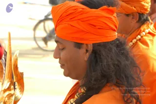 Outdoor photo in bright daylight showing a man in profile with eyes closed, wearing a saffron-orange turban and saffron-orange robes, with long dark wavy hair falling down his back and a beaded rudraksha-style necklace. Behind him a second man in orange robes and an orange turban with a beaded necklace stands in soft focus. At left edge a gold trident-shaped ceremonial staff is visible, and a blurred bicycle and roadway appear in the background.