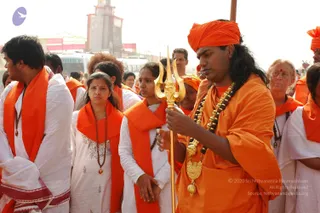 Outdoor photo of a religious procession at a crowded fairground. In the foreground a man in an orange turban and orange robe, with long dark hair and a dark-beaded rudraksha mala with gold ornaments, holds a tall golden trident (trishul) upright in both hands. Around him, devotees in white kurtas and white dhotis wear orange shawls; a woman in white with a mala stands beside a younger woman in white holding the same orange shawl. Behind them a red-and-white tower, parked buses and a crowd fill the background under bright sky.