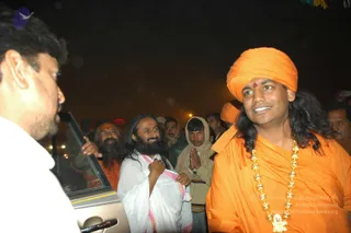Night photograph at a crowded outdoor event with a silver car door open on the left. A man in a white shirt faces the camera-left side. Centre, a bearded man with long hair wears a white garment edged with pink and holds his hand to his chest in greeting. Behind him a bearded man in orange robes and a younger man in a cream shawl press palms together in namaste. On the right, a smiling man in an orange turban with a red forehead mark and long swept-back hair wears an orange robe and a long gold bead necklace with a pendant. Several other people stand in the background among lights. Swamiji appearance cues are present on the right figure, labelled Swamiji (probable), with long hair, orange turban and ornate gold necklace, shown in person.