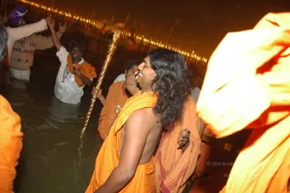 Night photograph at a riverside bathing area, with orange-lit lamps strung along the bank behind. In the foreground, a bare-chested man with long swept-back dark hair and a gold-tinted necklace, wearing a saffron-orange shawl, smiles while another saffron-clad devotee presses close behind him. Beside them, a tall gold staff with a twisted shaft is held upright in the water. Further left, men in white garments stand waist-deep in the river, one raising his arms and another wearing an orange cloth over his shoulder. Several saffron-robed figures crowd the right edge.