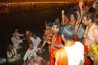 Night photograph at a river ghat during a Kumbh Mela bathing scene, with crowds standing in dark water and illuminated lights along the far bank. Several devotees in saffron and orange robes, some wet and wearing marigold and bead garlands, stand in the water with raised arms. A bald man in white stands at right with a rudraksha mala, and a long gold-topped staff with a silver finial rises beside the group. Two women in white kneel in the shallows, one smiling with hands together. A man in a white shirt and orange-red shawl raises his hand at left. Among the robed figures, one man with long dark hair and a crown-like headpiece in saffron cloth stands at center, though his identity cannot be determined from appearance.