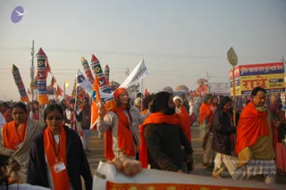 A large outdoor procession on a hazy winter morning at a Kumbh Mela-style gathering. Many devotees in saffron-orange shawls and scarves walk along a dusty road. A man in the centre wears a dark jacket over an orange shawl with a long beard and unruly hair, and a woman in a white jacket wears an orange headscarf and shawl while carrying a white banner. Colourful painted tall poles with cloth decorations rise behind them, and a white banner with blue lettering is held aloft. On the right, a man in an orange shawl holds a decorative ceremonial staff topped with a golden emblem beside a red and yellow sign reading police assistance and a TMT camp. Tents and more processional flags line the background.