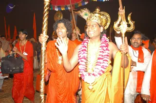 Night procession at the Allahabad Kumbh Mela with orange-clad devotees carrying a tall gold staff and a gold trident. The central figure, wearing a gold crown with a peacock-feather fan, a pink and white flower garland, a gold necklace and a yellow-gold cloth, raises a hand in blessing. Beside him a younger man in an orange robe with curly hair and a gold pendant chants. Behind them a gold-fringed multicoloured canopy and orange flags appear. On the right a man in white with an orange shawl and rudraksha mala holds a trident. The scene is lit by flash against a dark sky, with dry straw underfoot. Swamiji (probable): the central figure with long swept-back hair, a peacock-feather crown and ornate gold ceremonial dress, in a procession.