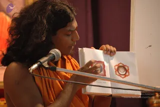 A long-haired man in an orange saffron shawl speaks into a microphone on a stand, holding open a book whose pages show red lotus chakra diagrams with a seed symbol at the centre. A small red tilak mark is on his forehead, and a maroon curtain stands behind him. The page shows a discourse setting with an open book being shown to an audience.