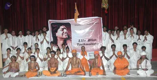 A large group of people, mostly in white clothing, pose with palms pressed together in namaste on a stage with red curtains. In the front row, several men wear saffron/orange robes and orange head cloths, some with rudraksha malas; one sits cross-legged holding a small brass lamp and a bell-like object on a white cloth. A banner reads Nithyananda Meditation Academy and Welcome to Life Bliss Technology, showing a portrait of a smiling man with long dark hair. An orange flag hangs at the top left and a decorative tassel hangs at the upper right.