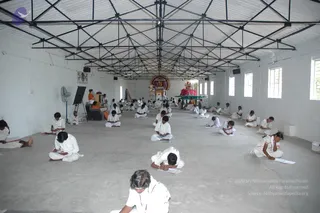 A photograph inside a bare, whitewashed hall with a steel truss roof and large windows. Many people in plain white clothes sit cross-legged on the concrete floor in rows, several writing on paper pads or sheets. At the far end, a decorated altar holds a deity murti with a pink and gold backdrop, a kalasha-style arrangement and garlands. A person in orange robes stands near a wooden stand at the left side of the room, and a whiteboard on an easel sits nearby. A ceiling fan and loudspeakers are visible.
