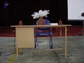 A photograph of an empty stage set-up at an indoor hall. A beige metal desk stands centre-stage with a blue chair wrapped in plastic and blue tape behind it. On the desk sit two small terracotta Ganesha figurines flanking a terracotta lattice vase holding white artificial flowers, plus a white box. A dark red curtain hangs behind, with a projection screen at the right edge and a cable running across the concrete floor. No people are visible.