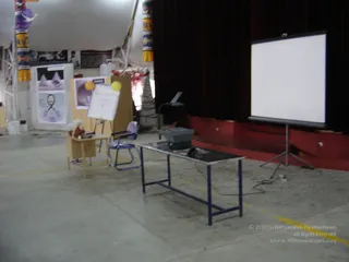 An empty indoor exhibition hall with a concrete floor and white tent ceiling. In the foreground stands a blue-framed metal table holding a projector, beside a blue-and-white chair and a small wooden side table with a stuffed toy. A tripod-mounted blank projection screen stands on a red-carpeted stage backed by dark red curtains. Left of centre, a wooden easel holds a whiteboard with two yellow balloons, and display boards show pyramid graphics and a framed portrait of a man with a printed label. Fabric-wrapped pillars with yellow and purple patterns frame the scene. No people are visible.