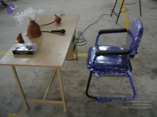 Photograph of an empty speaker setup in a large hall with a concrete floor. A light wooden table holds a woven terracotta vase with white artificial flowers, a small terracotta lamp with a red-tipped wick, a black corded microphone, a small brown figurine, and a dark hardcover book with a silver cover. Beside the table stands a blue upholstered armchair with black armrests, wrapped in clear plastic and bound with blue rope. Yellow floor markings and cables are visible. No people are present.